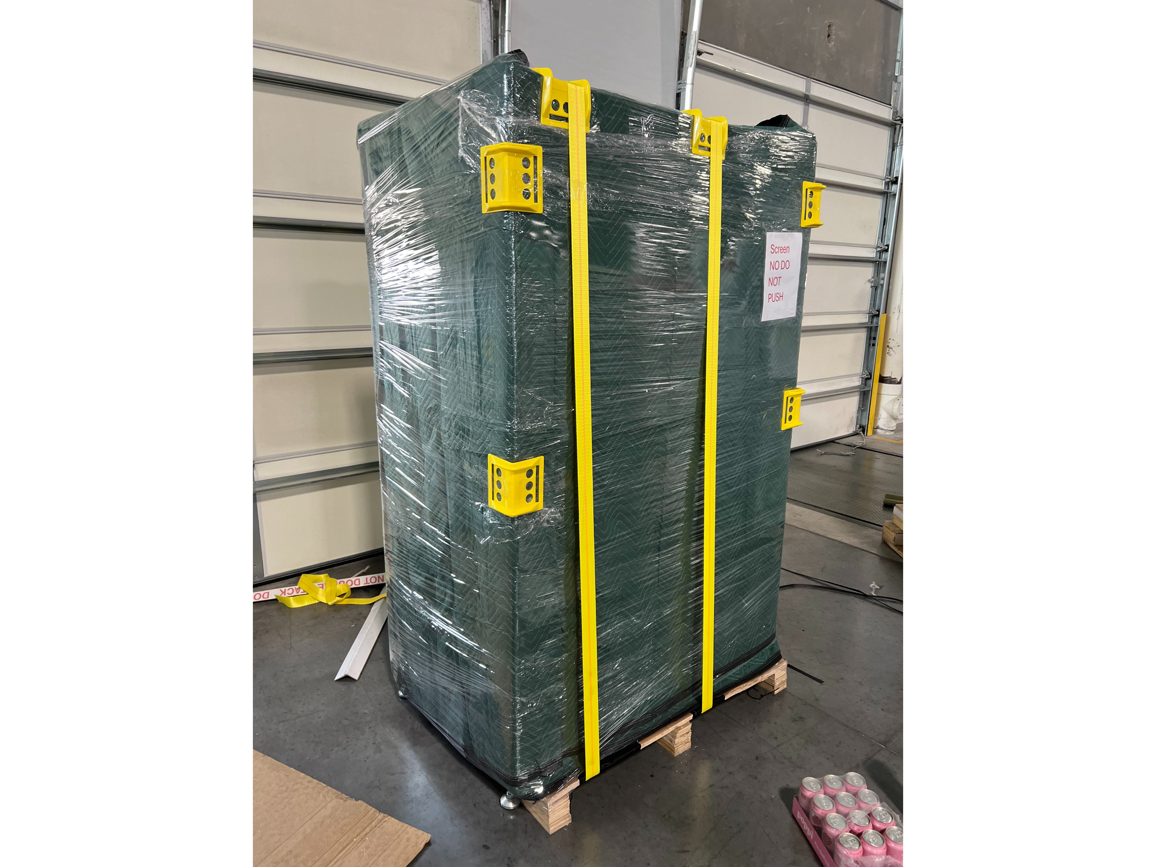 Vending machine securely packaged for transport with dark green protective blankets, clear shrink wrap, bright yellow straps, and yellow corner protectors on wooden pallet - professional moving service in Utah