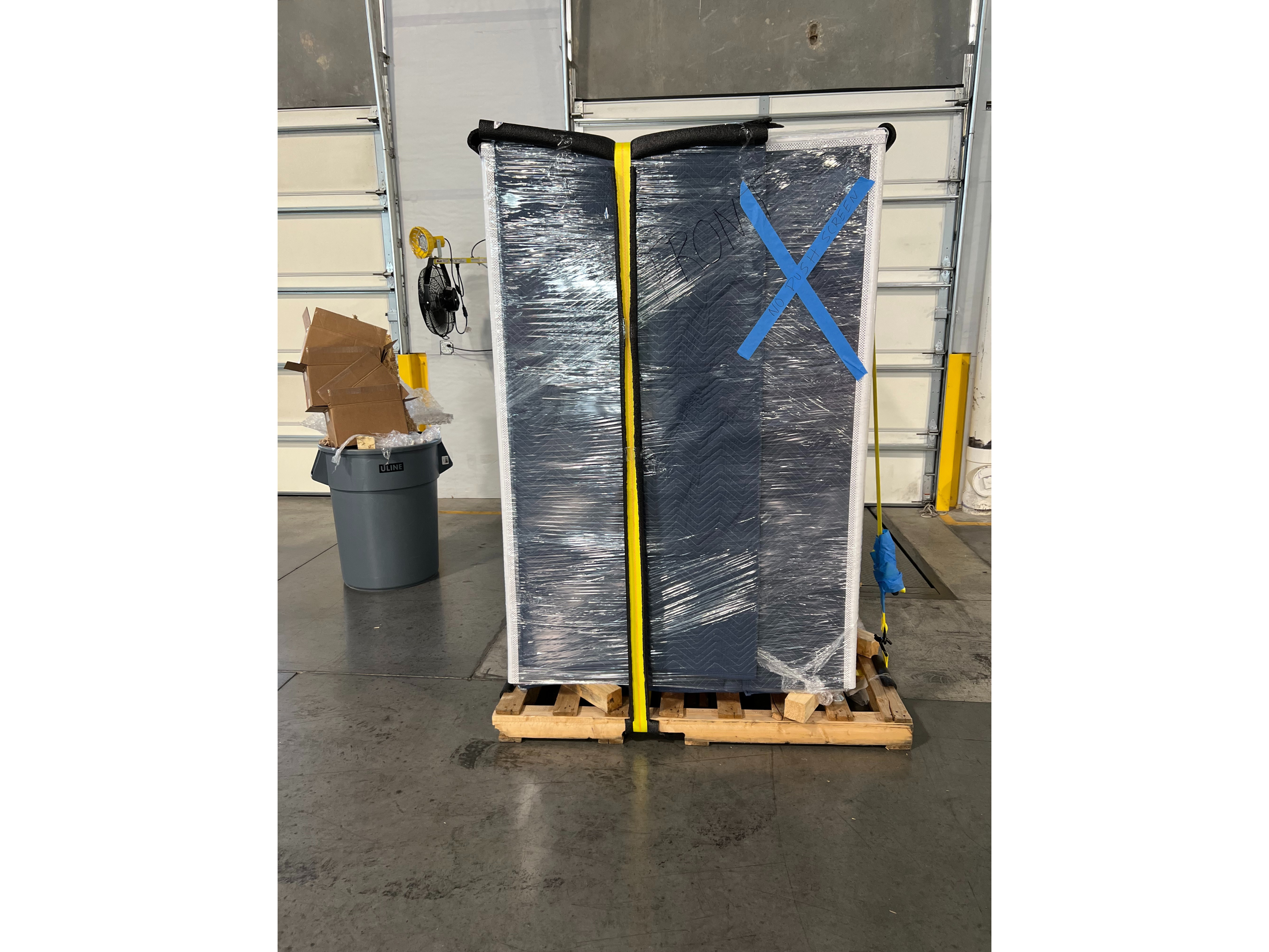 Vending machine wrapped in dark blue protective blankets and clear shrink wrap with yellow straps, foam padding, and safety markings secured on wooden pallets for safe transport